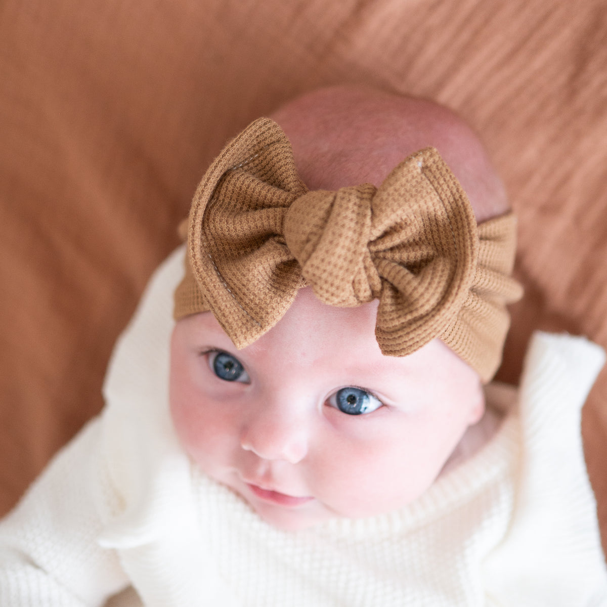 Top close-up view of a brown waffle knit baby headband, modeled on a baby with blue eyes and a white onesie.