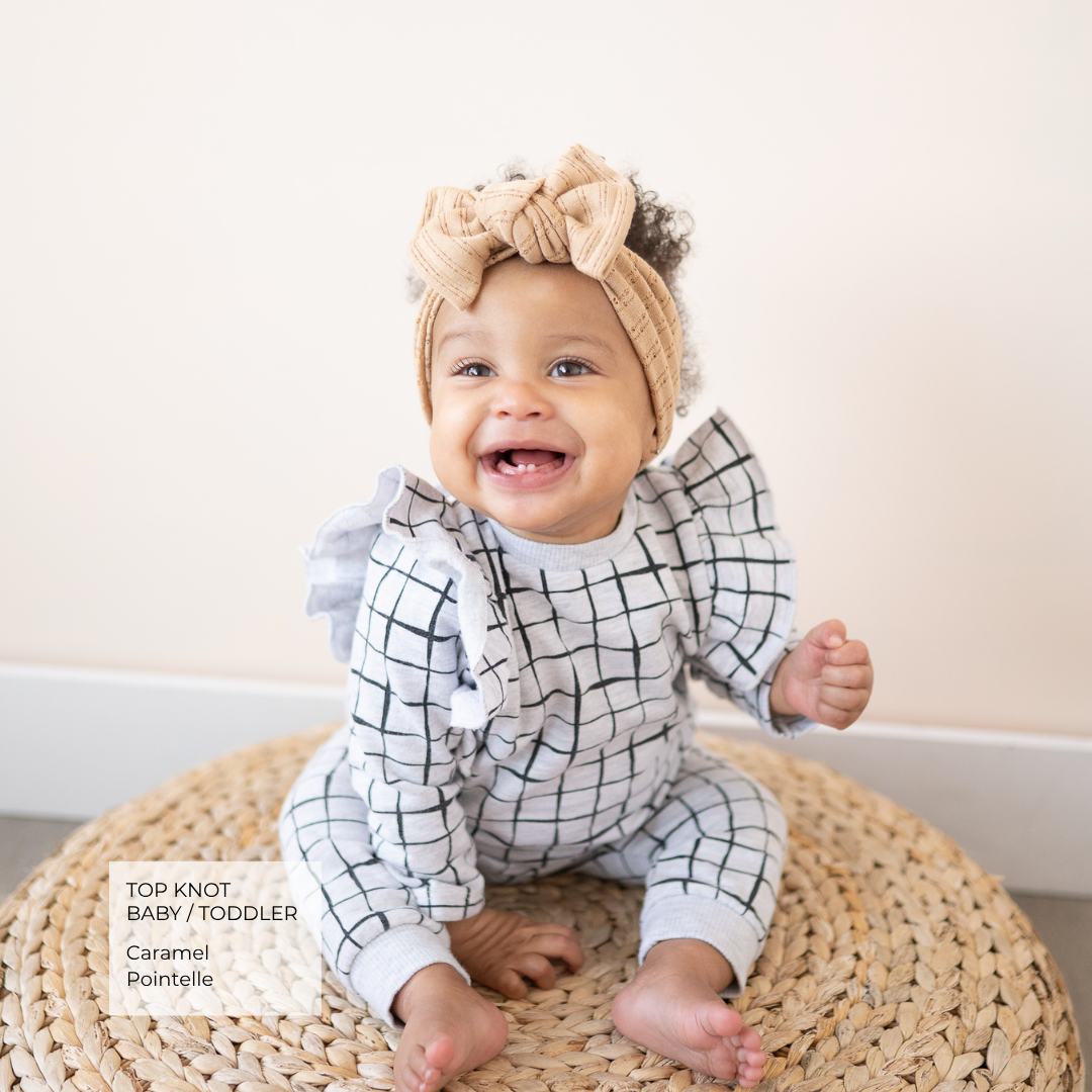 A baby wearing a caramel knit top knot headband and a black and white grid-patterned romper, sitting on a round woven straw rug against a light beige wall.