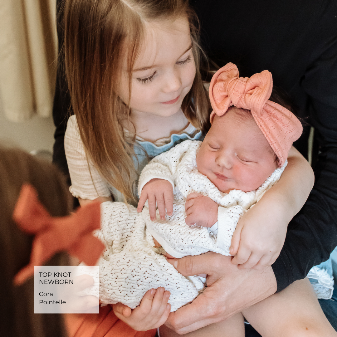 A newborn baby wrapped in a white knit blanket and wearing a coral pink top knot headband, being held by an adult while a young girl looks on. A text overlay in the bottom left reads 'TOP KNOT NEWBORN Coral Pointelle'.