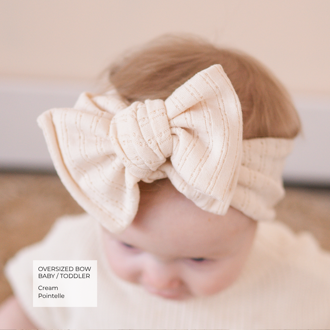 A baby wearing an oversized cream pointelle bow headband, shown from the back. A text overlay in the bottom left corner reads 'Oversized Bow Baby / Toddler Cream Pointelle'.