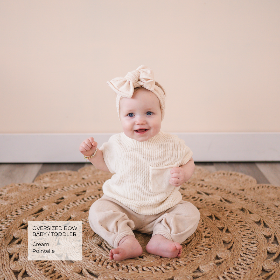 A baby sitting on a woven jute rug wearing a cream ribbed knit romper with a chest pocket and a matching oversized bow headband. A text overlay in the bottom left reads 'Oversized Bow Baby / Toddler Cream Pointelle'.