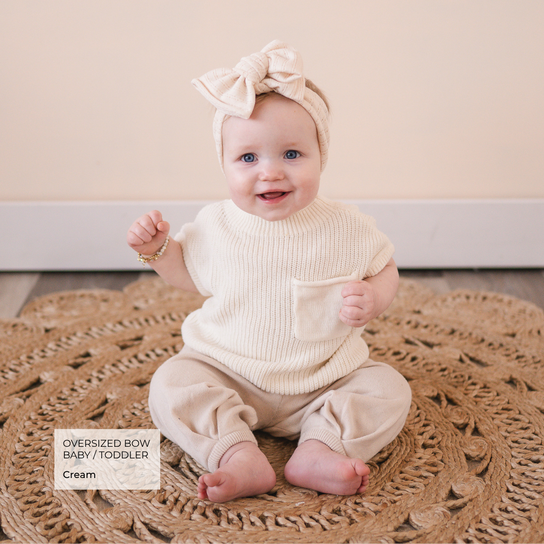 Baby wearing a cream outfit with an oversized bow headband on a textured rug.