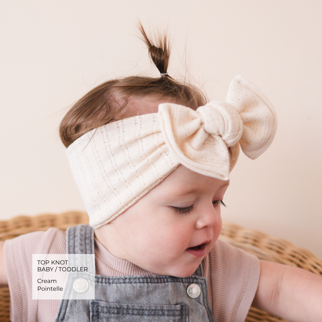 A baby wearing a cream cotton top knot headband with a large bow, styled over a pink ribbed top and denim overalls, sitting in a wicker basket.