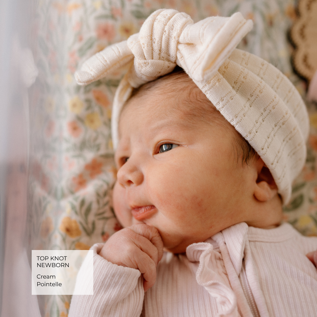 A close-up of a newborn baby wearing a cream cotton pointelle knit top knot headband with a bow, paired with a pale pink ribbed romper. The baby is resting against a soft floral-patterned fabric background.