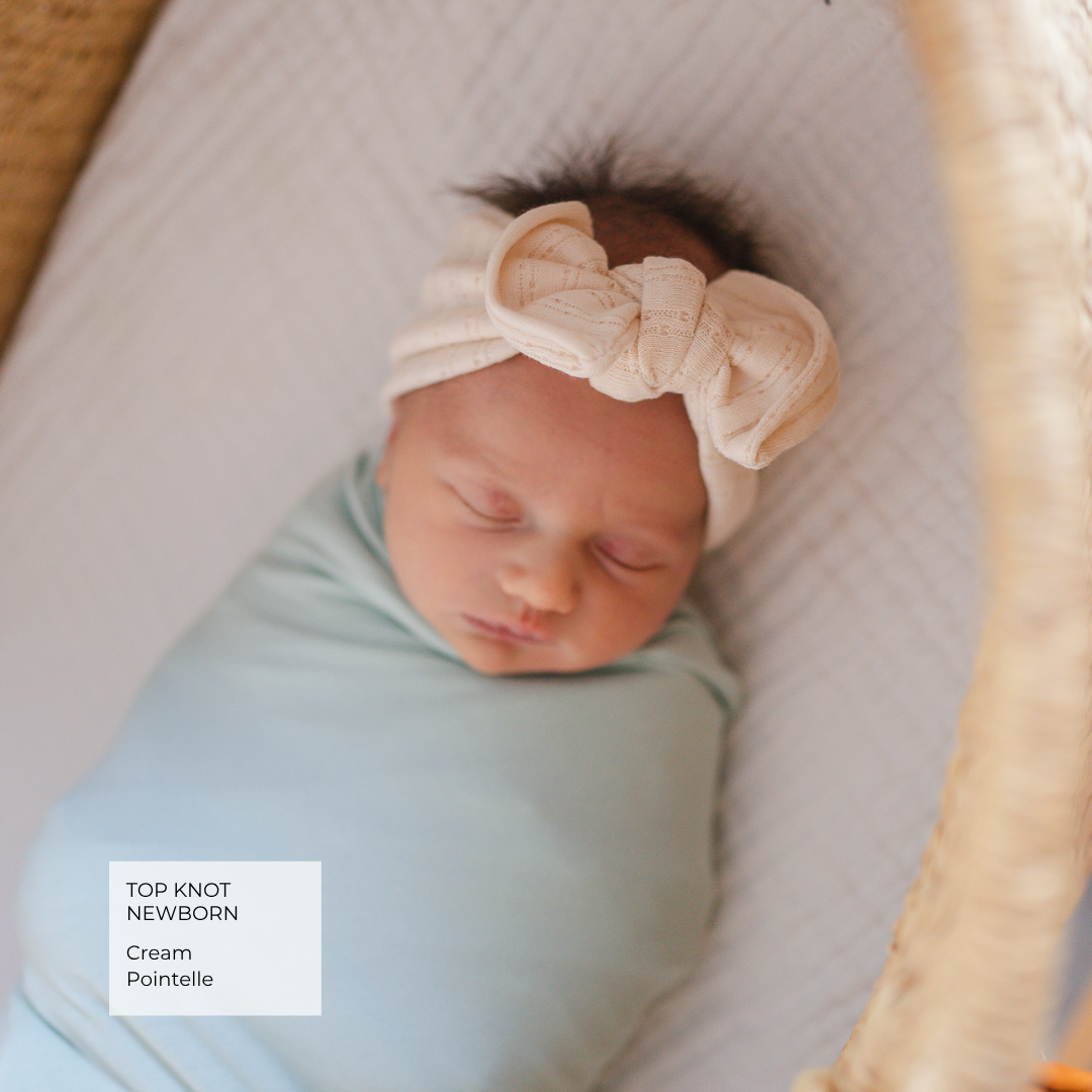 A sleeping newborn baby swaddled in a pale blue blanket and wearing a cream knit top knot headband, resting inside a woven bassinet. A text label in the bottom left reads 'TOP KNOT NEWBORN Cream Pointelle'.