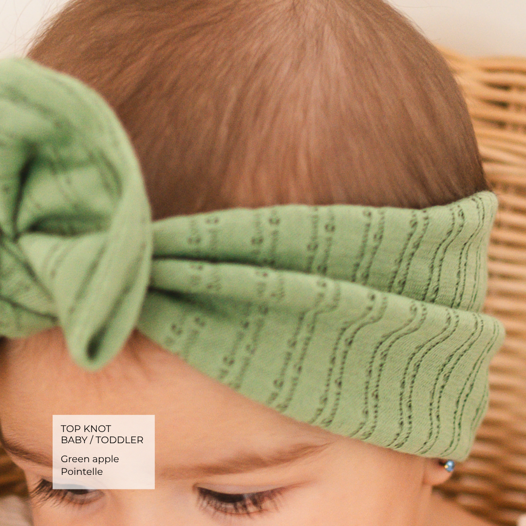 A close-up of a baby wearing a sage green top knot headband with a textured wavy knit pattern. A text label in the bottom left corner reads 'TOP KNOT BABY / TODDLER Green apple Pointelle'.