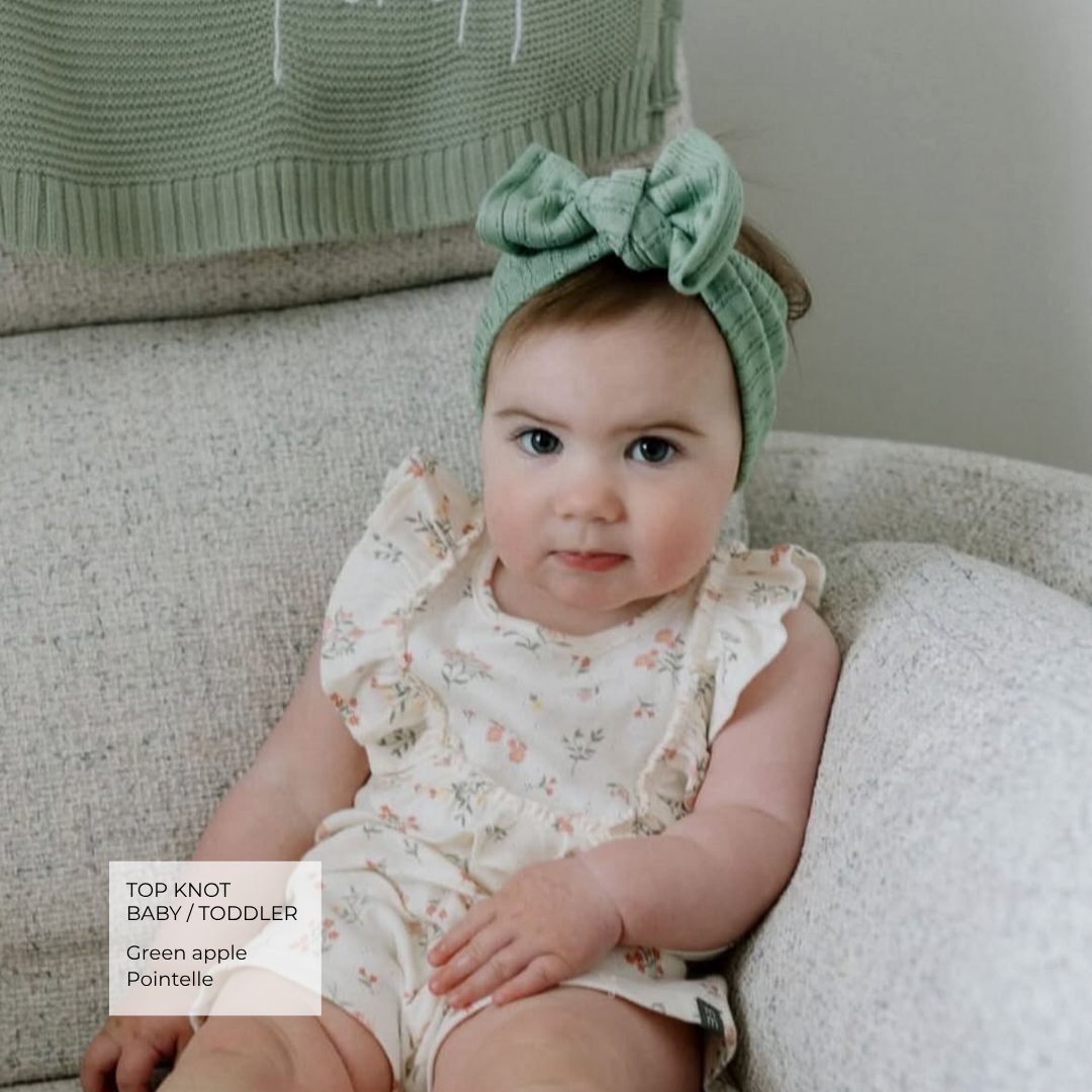 A baby wearing a green knitted top knot headband with a large bow and a cream floral romper, sitting on a textured light gray sofa. A text overlay in the bottom left reads 'TOP KNOT BABY / TODDLER Green apple Pointelle'.