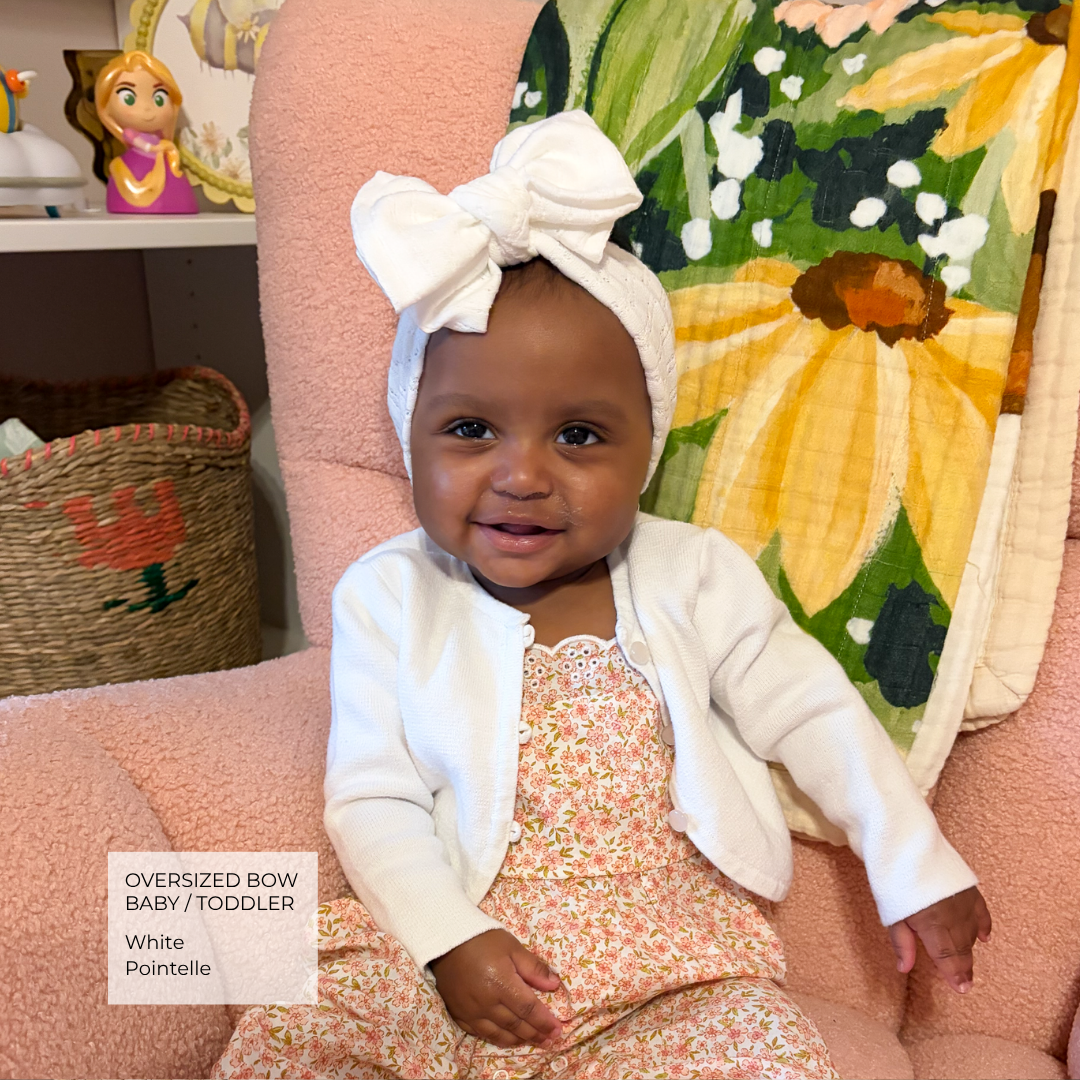 A baby wearing an oversized white bow headband and a white cardigan over a floral dress, sitting on a pink chair in a home setting.