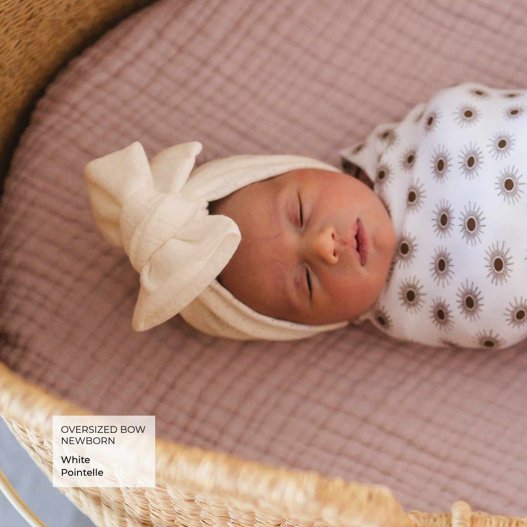 A sleeping newborn baby wrapped in a white swaddle with brown sunburst patterns, resting in a woven basket on a pink muslin mattress. The baby is also wrapped in a cream-colored bow headband.