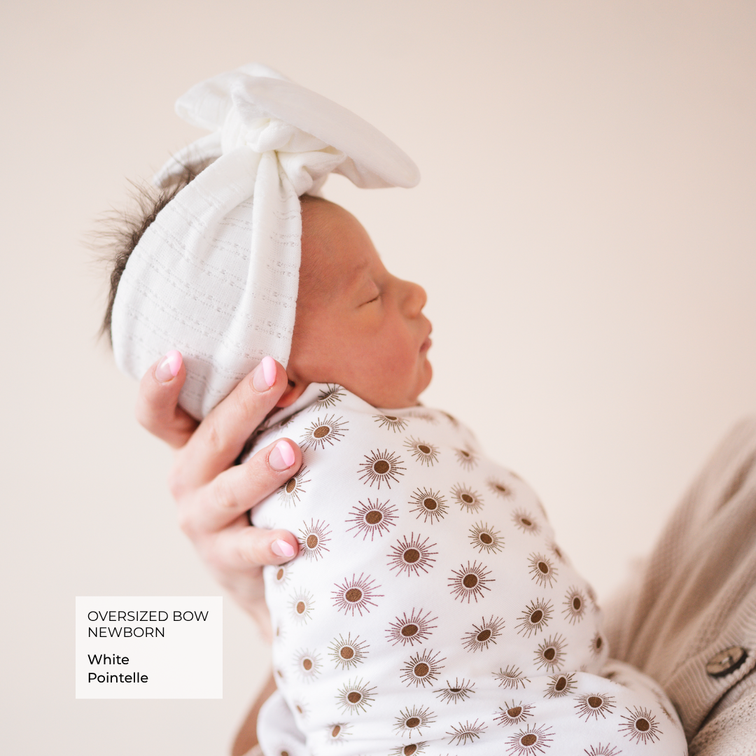 A newborn baby wearing an oversized white bow headband and a white swaddle blanket with a brown sunflower print, held by an adult hand against a soft beige background.