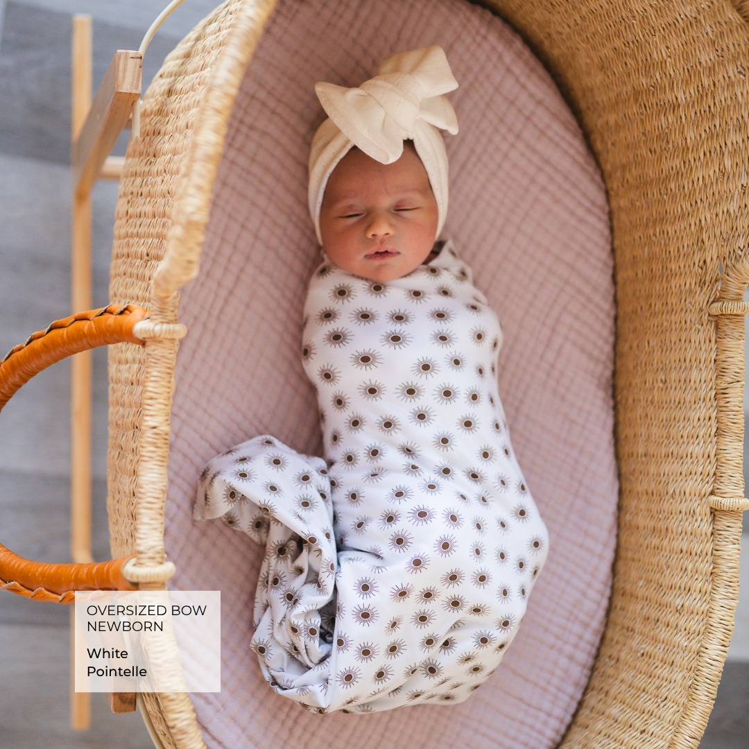 A newborn baby is sleeping peacefully inside a woven wicker Moses basket. The baby is swaddled in a white muslin blanket with a brown sunflower pattern and wears an oversized cream bow bonnet.