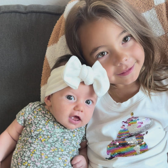Older sister sitting with infant baby girl wearing a white waffle top knot bow and smiling.