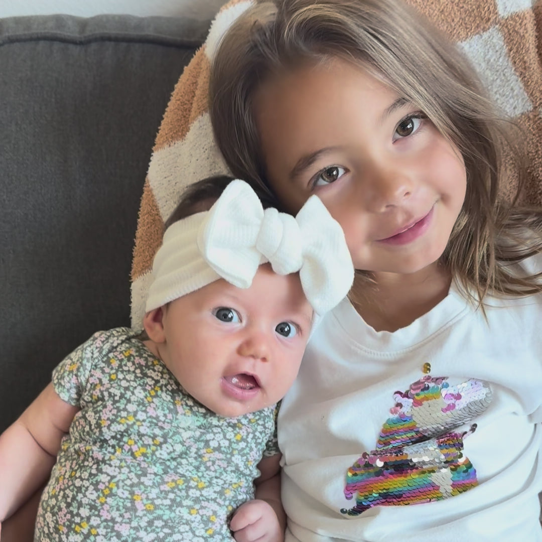 Older sister sitting with infant baby girl wearing a white waffle top knot bow and smiling.