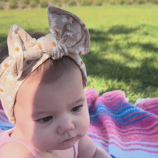 Baby wearing taupe daisy print top knot headband, playing on a bright colored blanket in the grass.