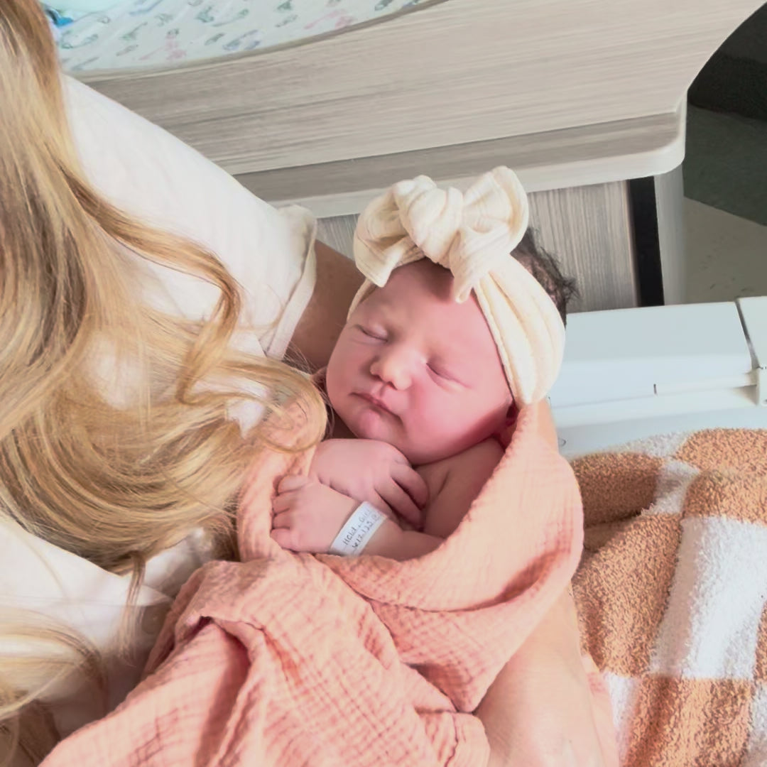 Newborn baby girl in hospital wearing first bow, a cream pointelle top knot. Mother is holding the baby in coral swaddle.
