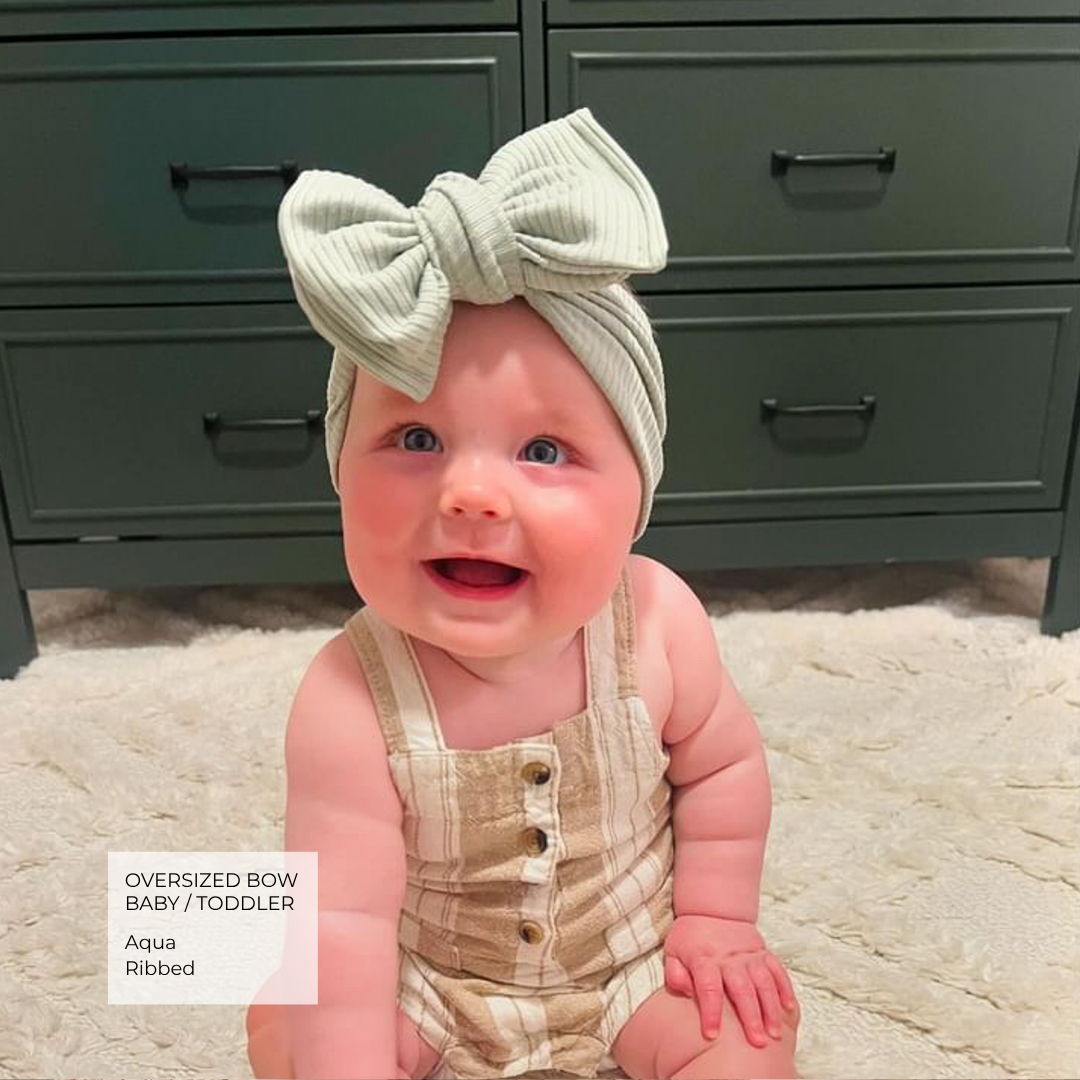 A smiling baby toddler wearing an oversized ribbed bow headband in aqua and a beige-and-cream striped romper, sitting on a cream rug in front of a dark green dresser.