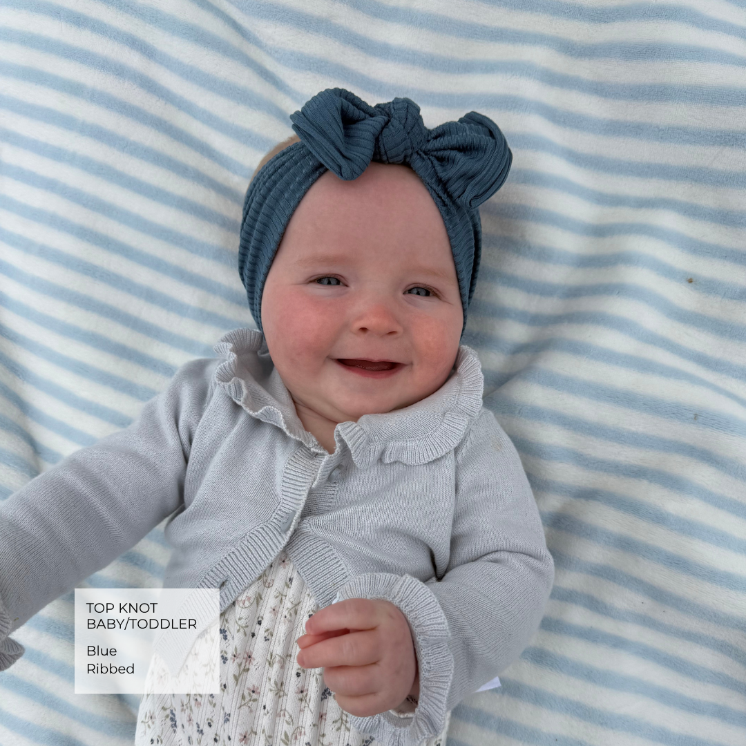 A baby wearing a blue ribbed top knot headband and a light gray cardigan, lying on a blue and white striped blanket. A text overlay in the bottom left corner reads 'TOP KNOT BABY/TODDLER Blue Ribbed'.