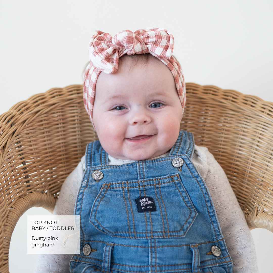 A baby wearing a dusty pink and white gingham top knot headband with a large bow, paired with blue denim overalls, sitting in a wicker chair. A text overlay in the bottom left corner reads 'TOP KNOT BAB / TODDLER Dusty pink gingham'.
