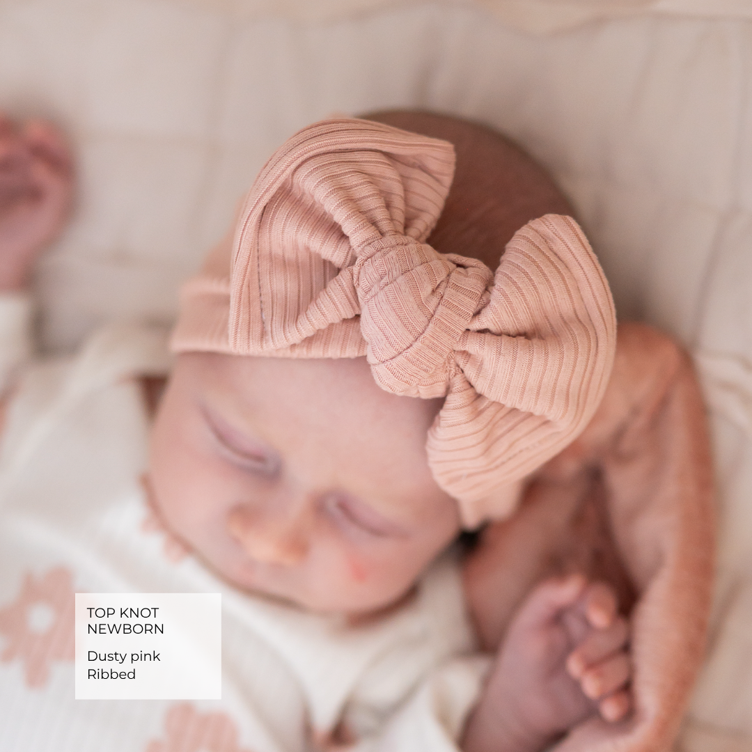 A sleeping newborn baby wearing a dusty pink ribbed top knot headband with a large bow, lying on a soft cream blanket. A text overlay in the bottom left reads 'TOP KNOT NEWBORN Dusty pink Ribbed'.