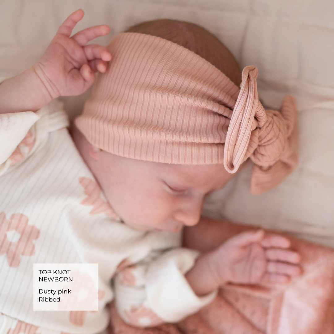 A newborn baby wearing a dusty pink ribbed top-knot headband with a bow, lying on a soft cream blanket. A text overlay in the bottom left reads 'TOP KNOT NEWBORN Dusty pink Ribbed'.
