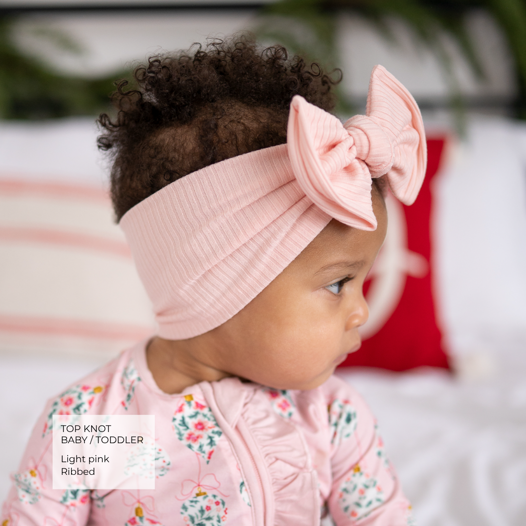 A baby wearing a light pink ribbed top knot headband with a large bow, paired with a matching pink floral long-sleeve romper. The photo is taken in a soft-focus indoor setting with a white pillow and red accent in the background.