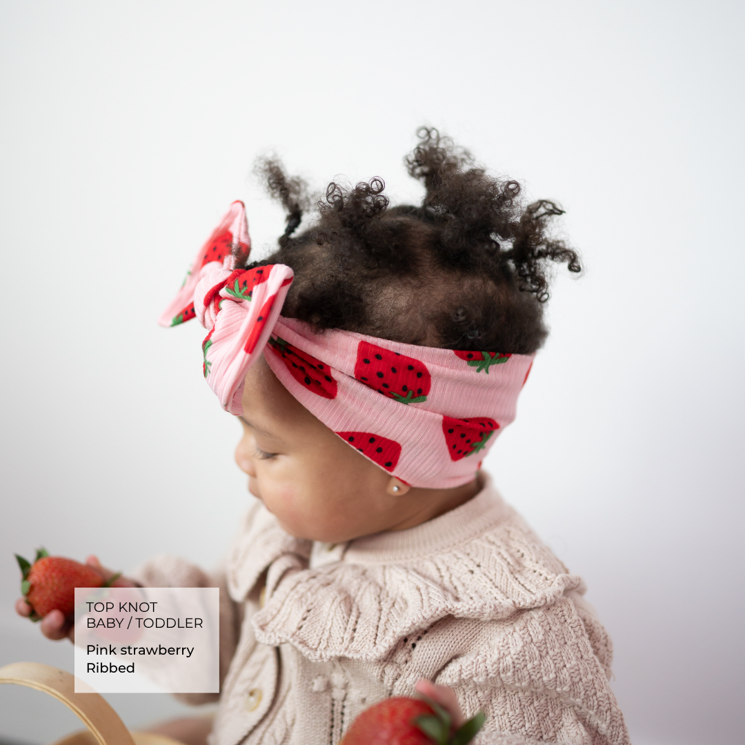A toddler wearing a pink ribbed knit top and a matching pink strawberry-patterned top knot headband, holding strawberries against a plain light background.