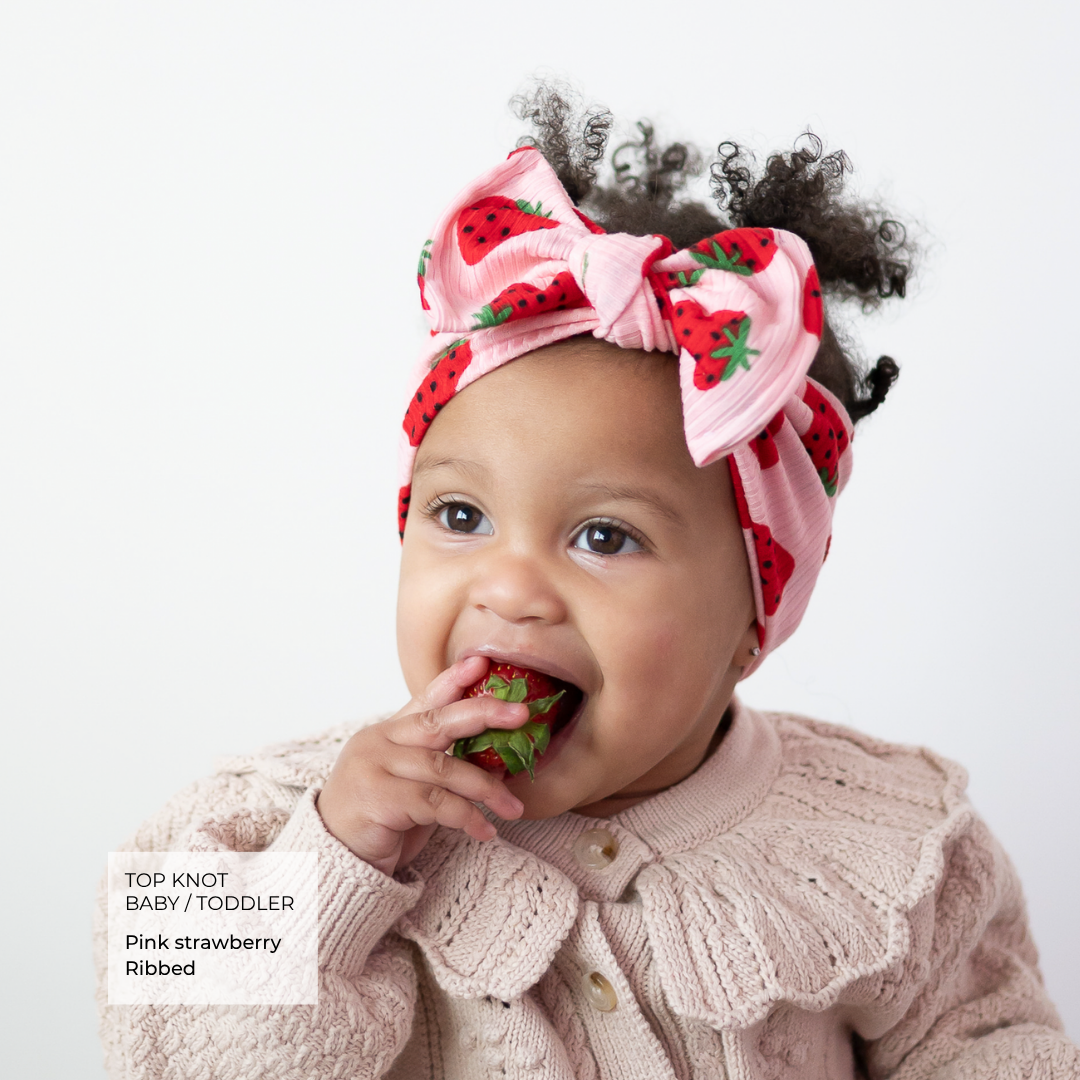 A baby wearing a pink strawberry-print top knot headband and a matching pink knit cardigan, eating a strawberry against a plain light background. A text box in the lower left reads 'TOP KNOT BABY / TODDLER Pink strawberry Ribbed'.