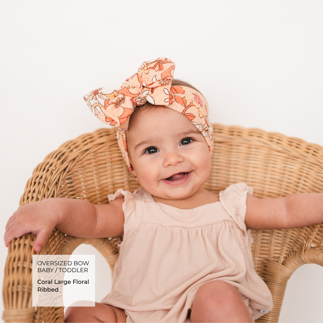 A baby wearing an oversized coral pink floral bow headband and a light beige ribbed romper, sitting in a wicker chair against a white background. A text overlay in the bottom left reads 'OVERSIZED BOW BABY / TODDLER Coral Large Floral Ribbed'.