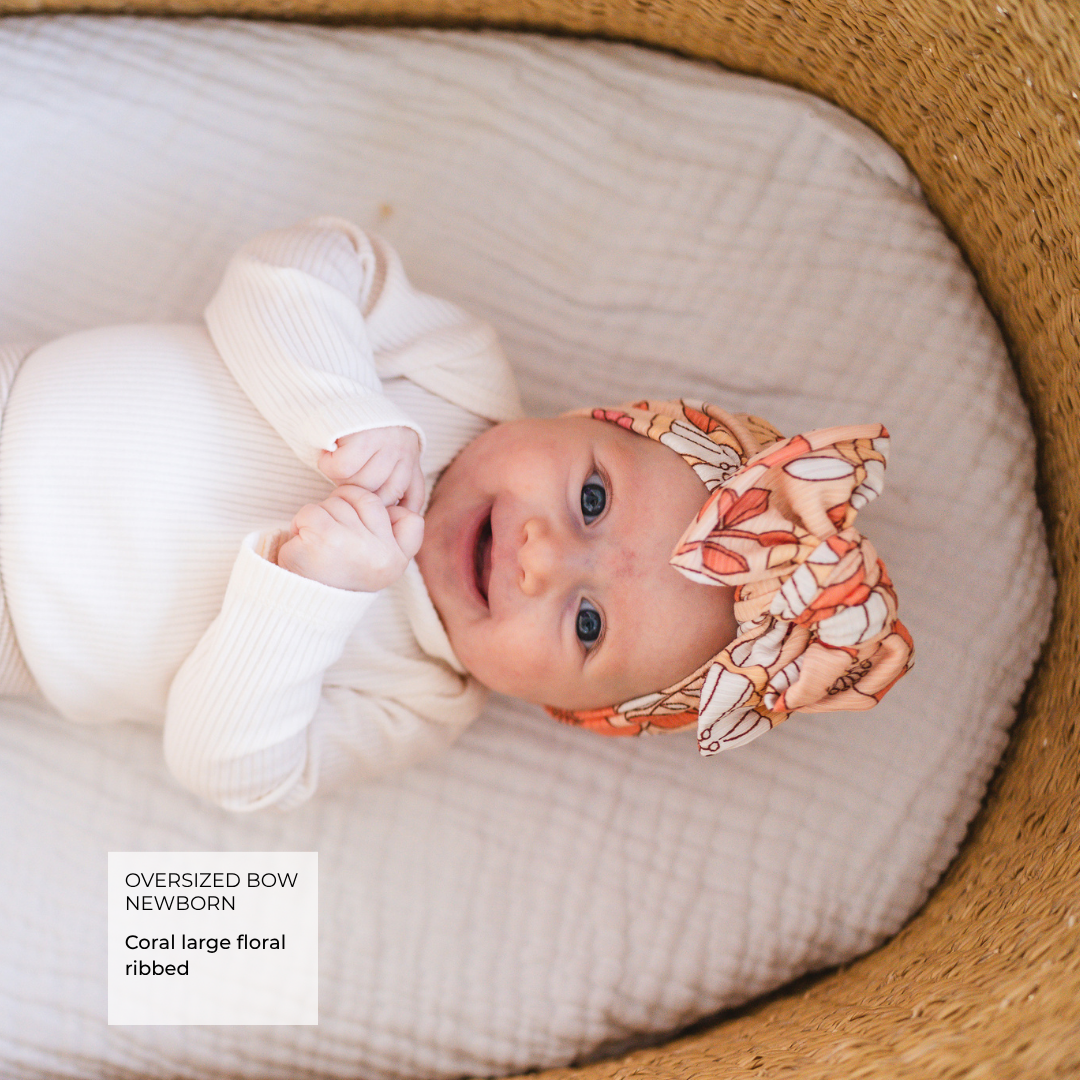 A newborn baby wearing a white ribbed bodysuit and a coral floral oversized bow headband, lying on a textured cream blanket inside a woven basket. A text overlay in the bottom left reads 'Oversized Bow Newborn Coral large floral ribbed'.