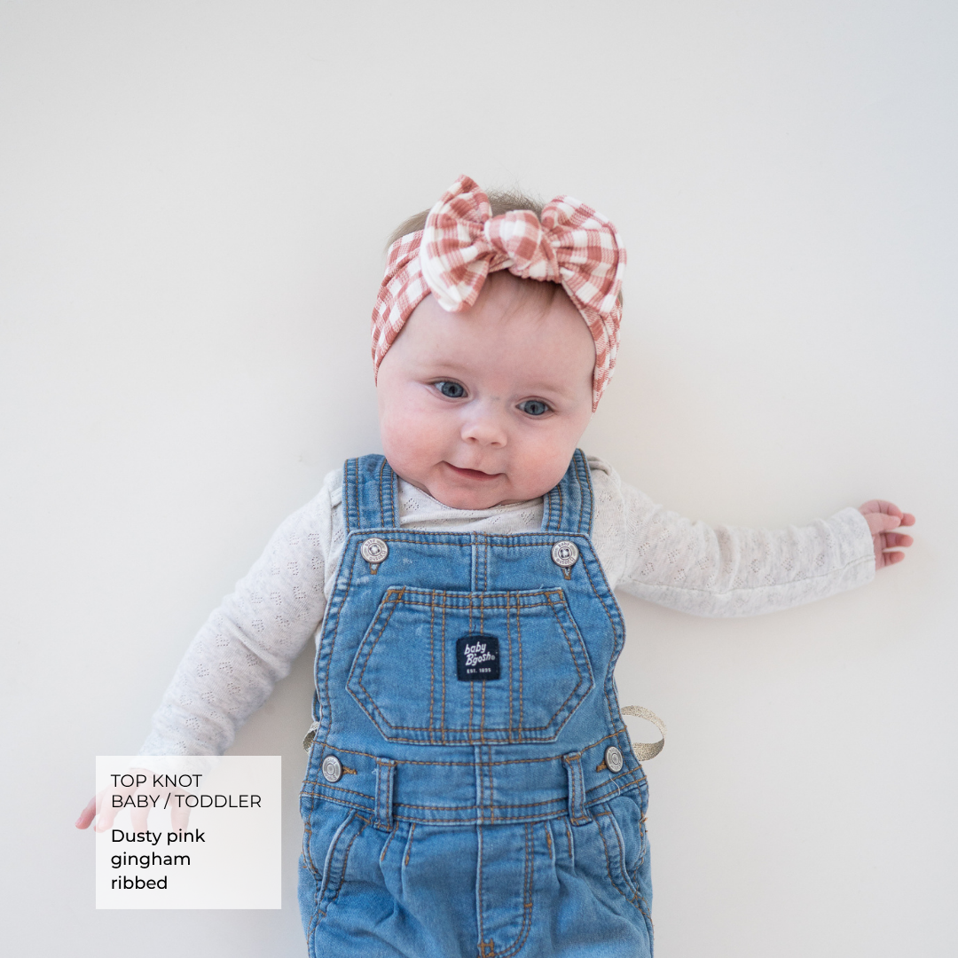 A baby wearing a dusty pink gingham bow headband and matching denim overalls over a white long-sleeve shirt, lying on a light off-white background. A text box in the bottom left corner reads 'TOP KNOT BABY/TODDLER Dusty pink gingham ribbed'.