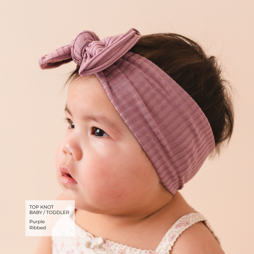 A close-up of a baby wearing a dusty purple ribbed fabric headband with a top knot and bow detail. The baby is shown in profile against a solid pale peach background, with a text overlay in the bottom-left corner reading 'TOP KNOT BABY / TODDLER Purple Ribbed'.