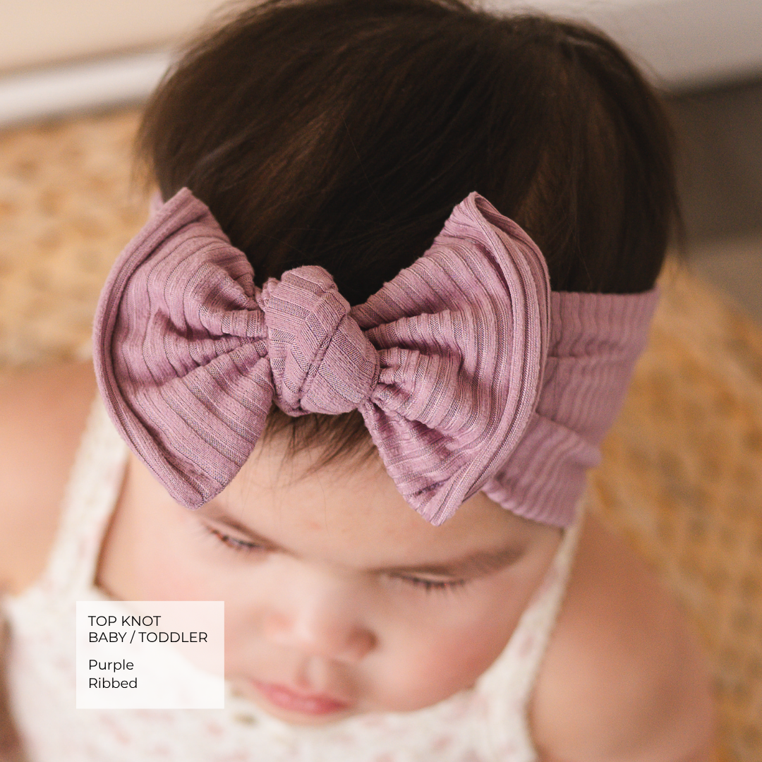 Baby wearing a large purple bow headband with blurred background