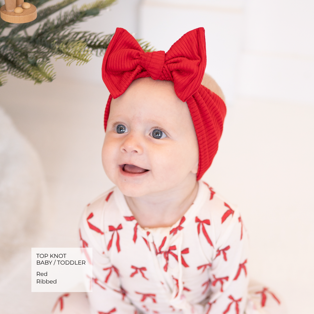 A baby wearing a red ribbed corduroy top knot headband with a large bow, paired with a white top featuring a red bow pattern. A text overlay in the bottom left corner reads 'TOP KNOT BABY / TODDLER Red Ribbed'.