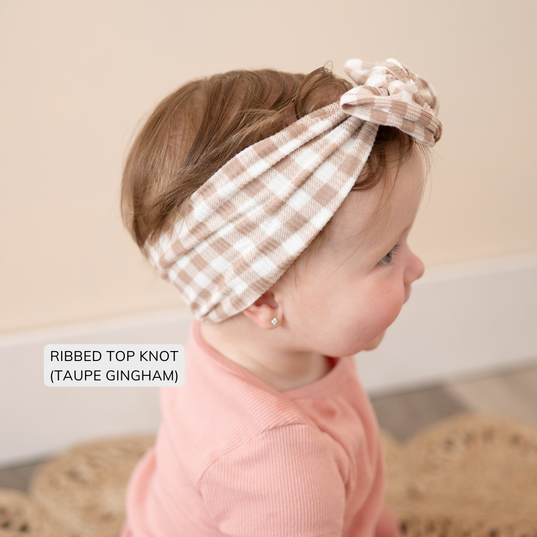 Child wearing a ribbed top knot headband with beige and white gingham pattern, sitting on a textured surface.