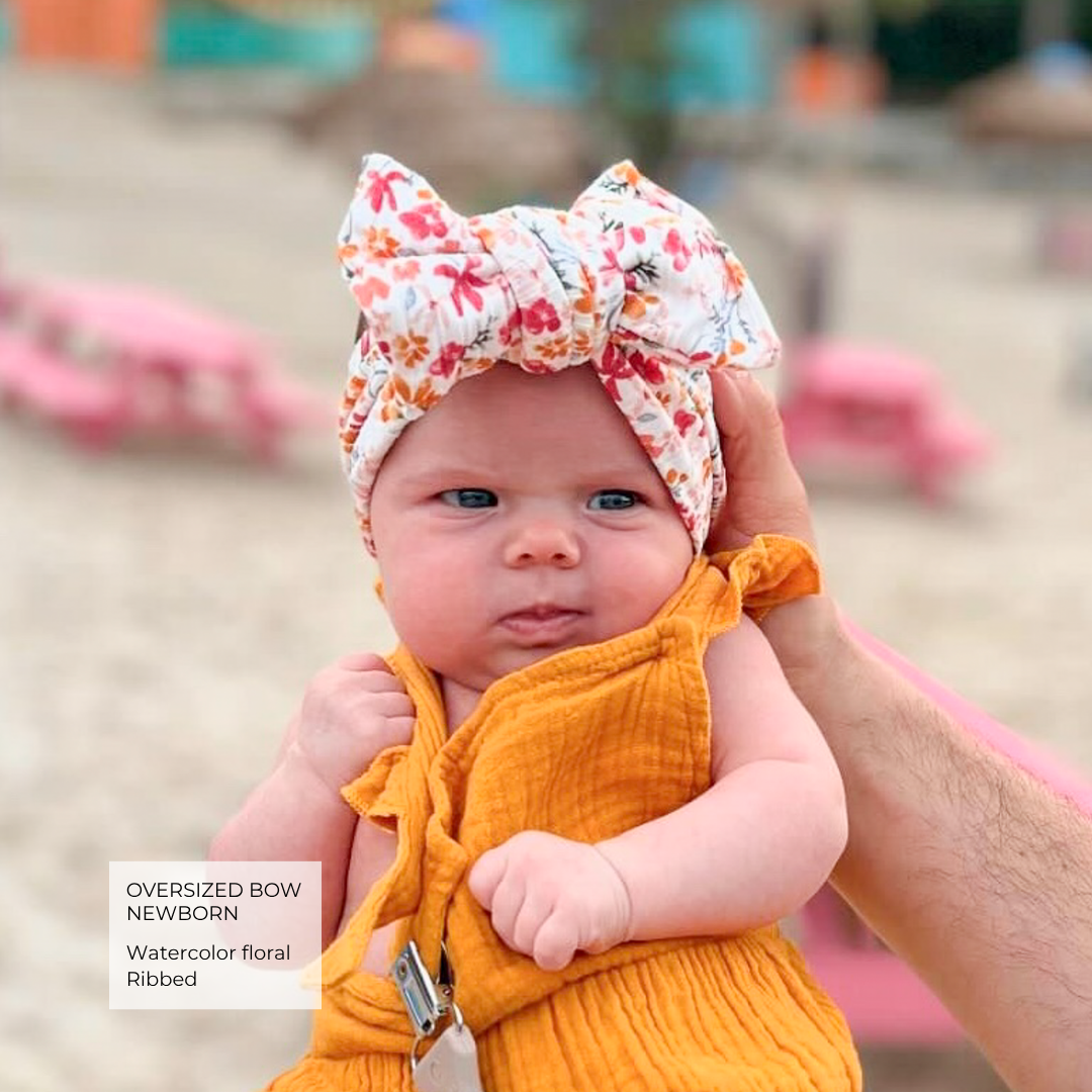 A baby wearing a mustard yellow ribbed romper and a white bow headband with a watercolor floral print, held up by an adult hand on a beach background.