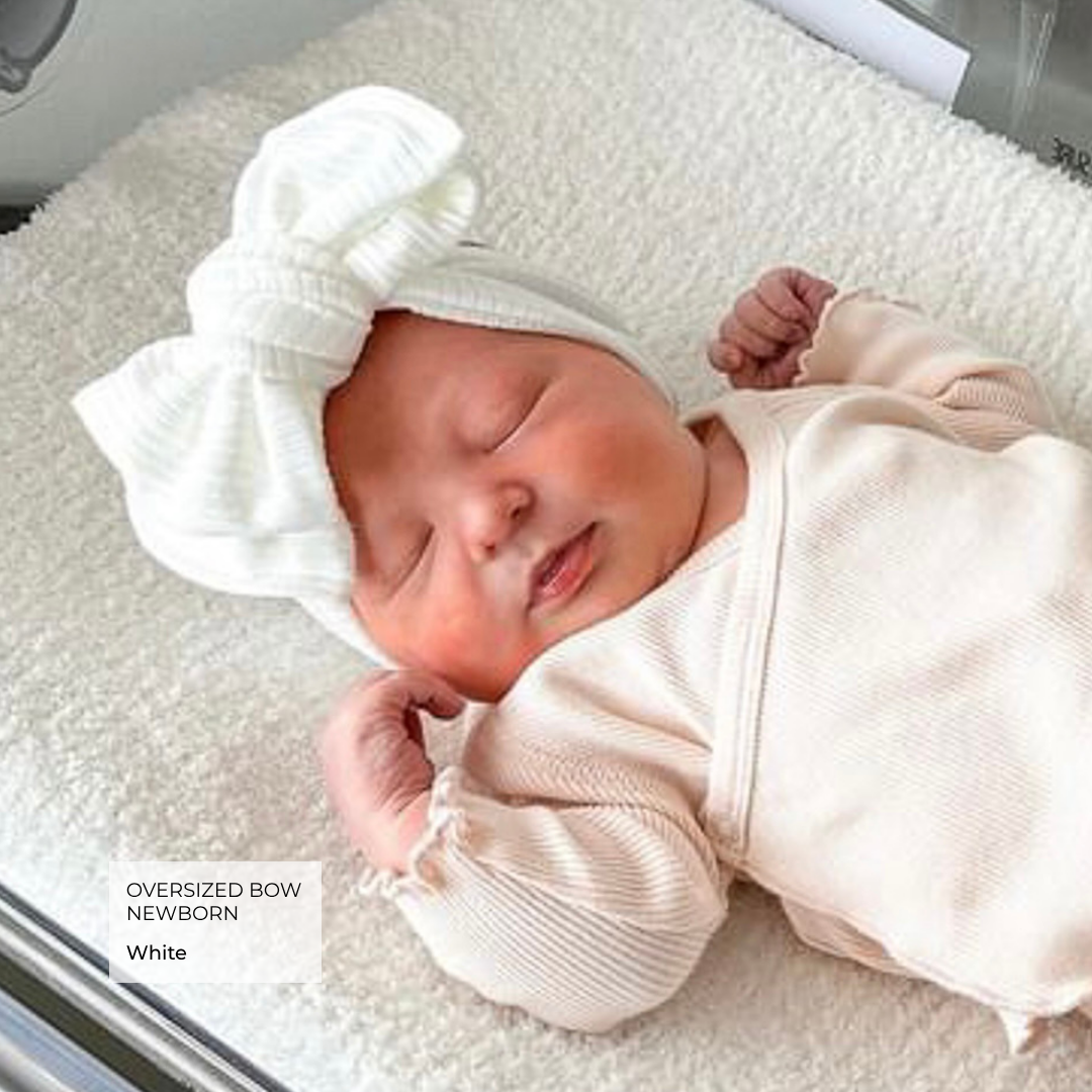 Newborn baby wearing a white oversized bow headband and pink outfit on a light surface.