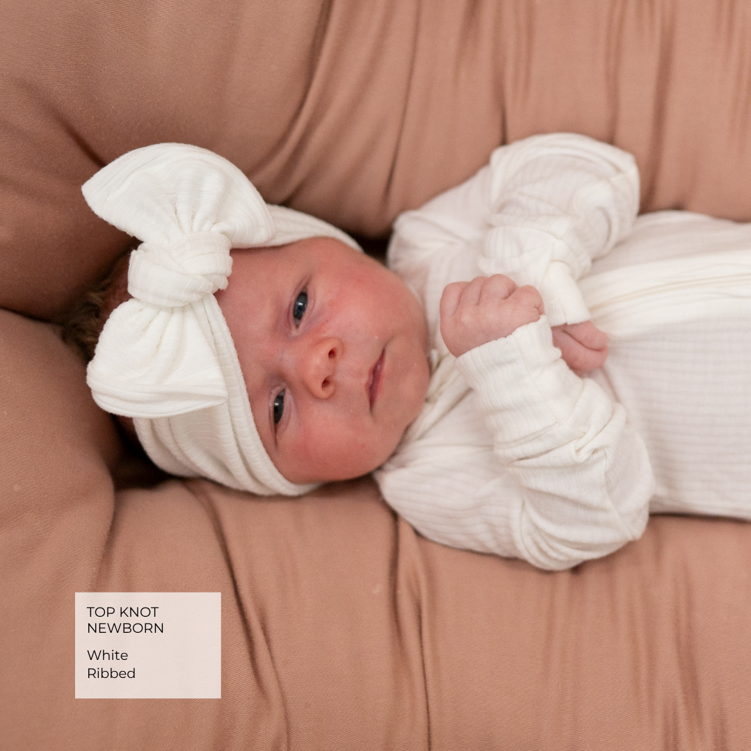 A newborn baby wearing a white ribbed top-knot bonnet and matching white ribbed bodysuit, lying on a soft brown blanket. A text label in the bottom-left corner reads 'TOP KNOT NEWBORN White Ribbed'.