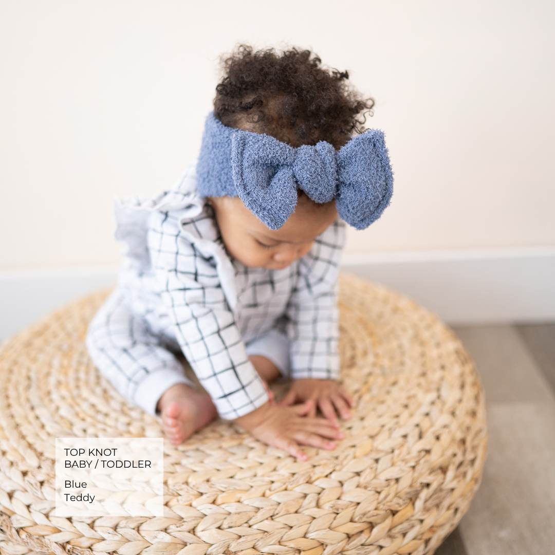 A baby wearing a blue textured top knot bow headband and a white and black checkered romper, sitting on a round woven straw stool against a light beige wall.