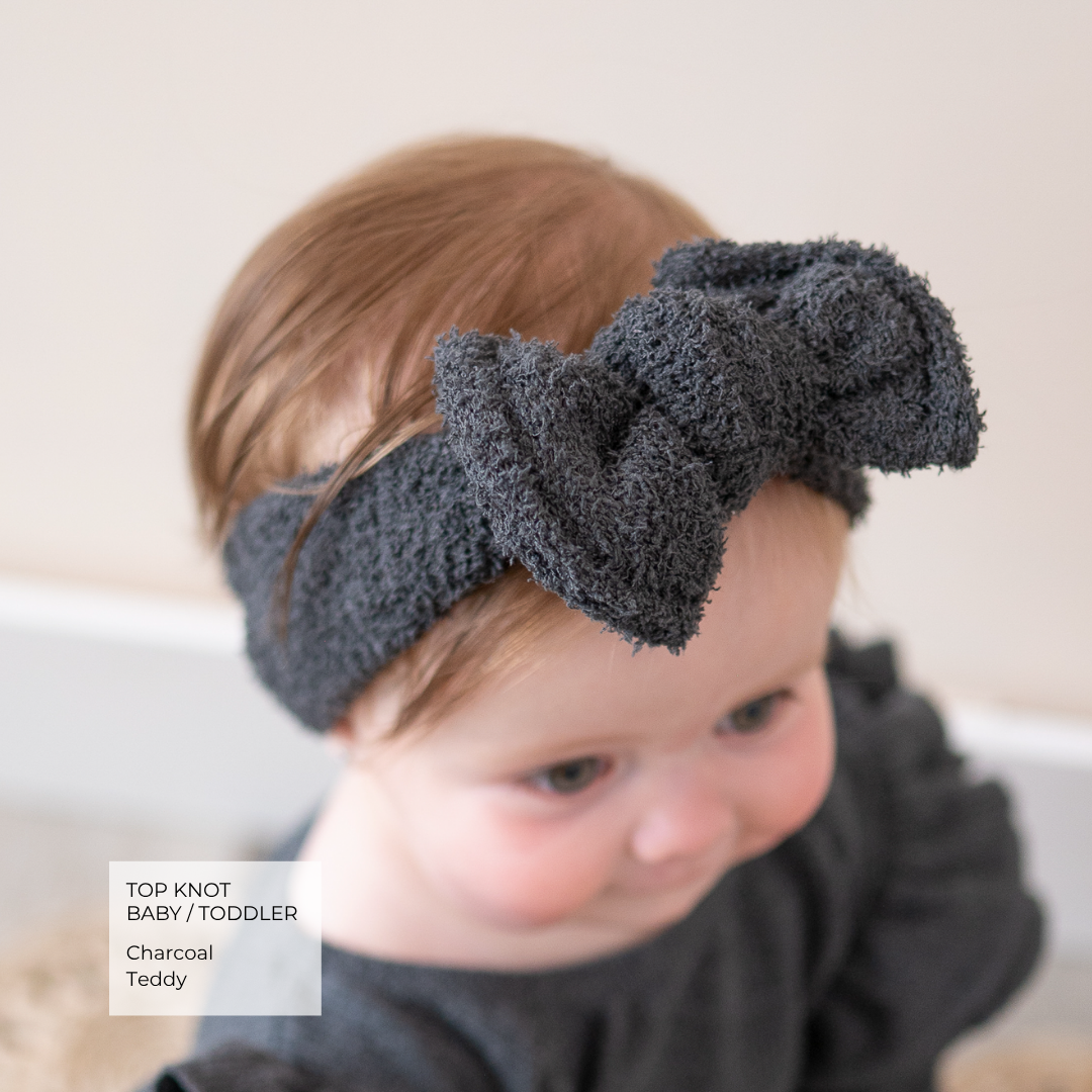 A baby wearing a dark charcoal gray textured top knot headband with a large bow, shown in a close-up portrait. A text overlay in the bottom left reads 'TOP KNOT BAB / TODDLER Charcoal Teddy'.