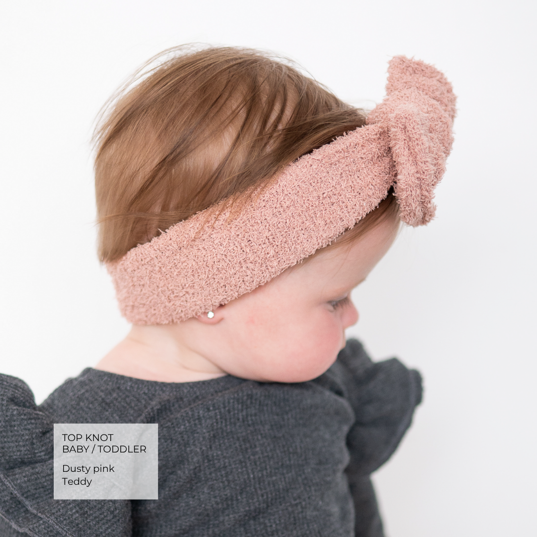 A toddler wearing a dusty pink textured top-knot headband, shown from the side against a plain white background. A text overlay in the bottom-left corner reads 'TOP KNOT BABY / TODDLER', 'Dusty pink', and 'Teddy'.