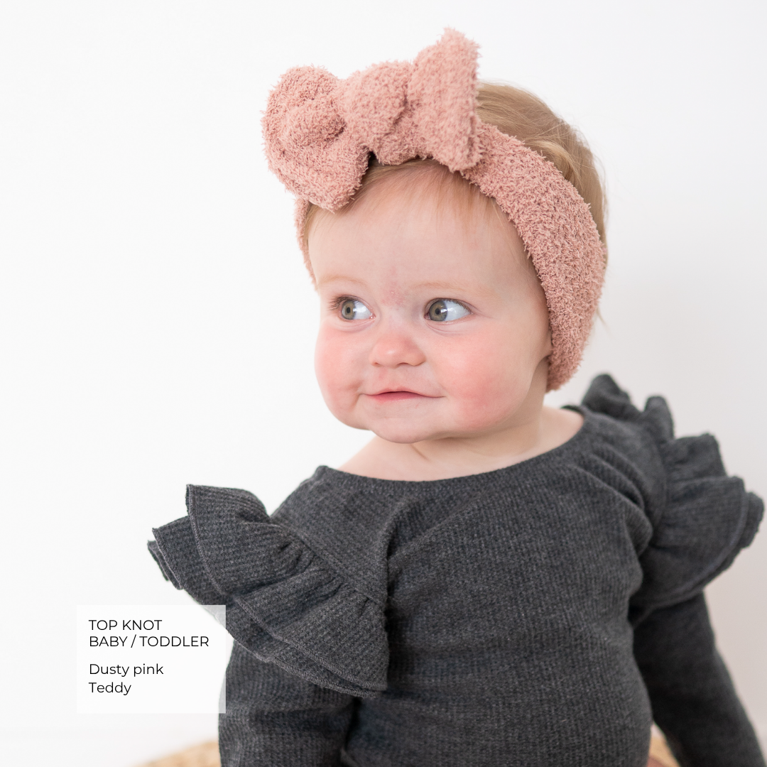 A baby wearing a dusty pink textured top-knot headband and a dark gray ruffled long-sleeve top, photographed against a clean white background with a text label in the lower left.