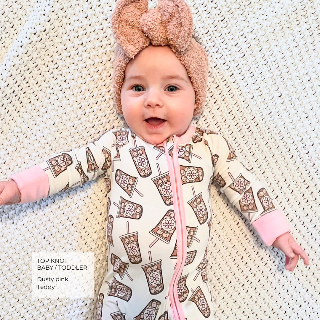 A baby lying on a white textured blanket wearing a dusty pink top-knot headband and a matching footless one-piece sleeper with a brown and white pattern and pink zipper.