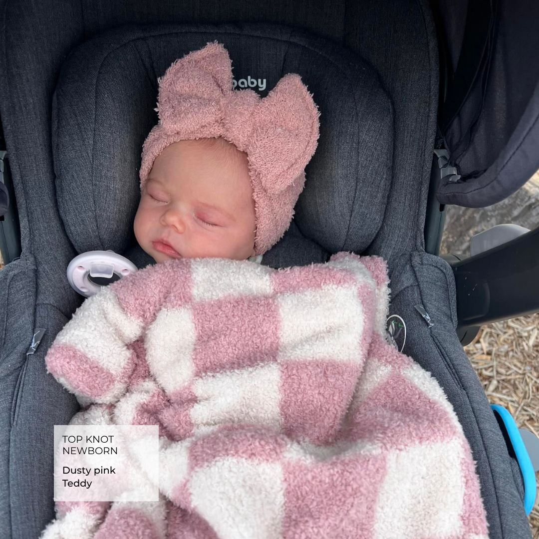 A sleeping newborn baby wearing a dusty pink knit hat with a large bow and wrapped in a pink and white checkered fuzzy blanket, resting in a dark gray stroller. A text overlay in the bottom left reads 'TOP KNOT NEWBORN Dusty pink'.