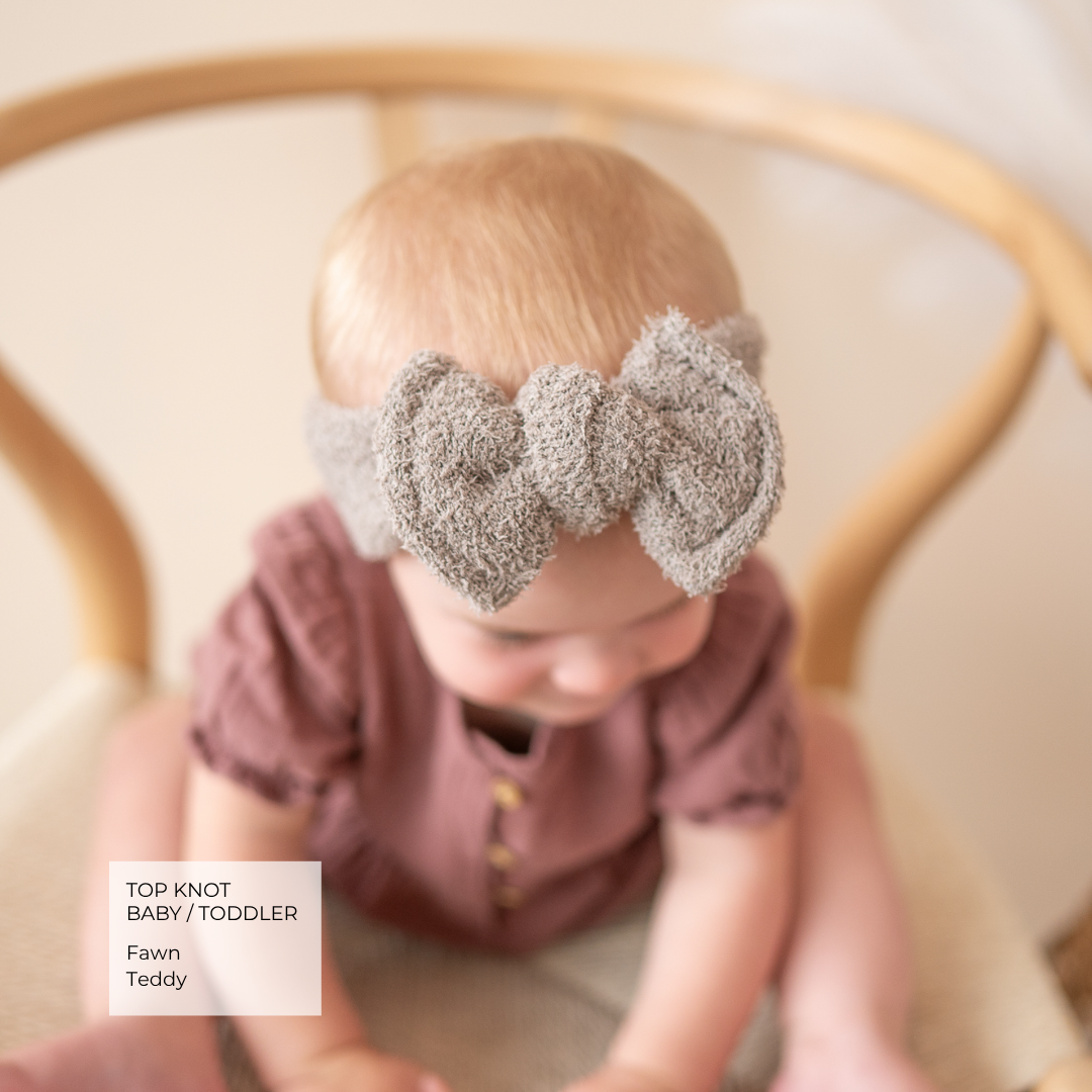 A baby wearing a textured gray top-knot bow headband, sitting in a wooden chair. The text overlay in the bottom left reads 'TOP KNOT BABY / TODDLER Fawn Teddy'.