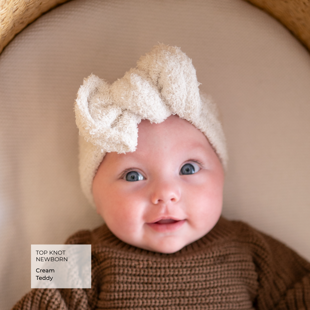 A close-up of a newborn baby wearing a cream-colored textured top knot headband and a brown knit sweater, set against a soft beige background with a wooden cradle edge visible.