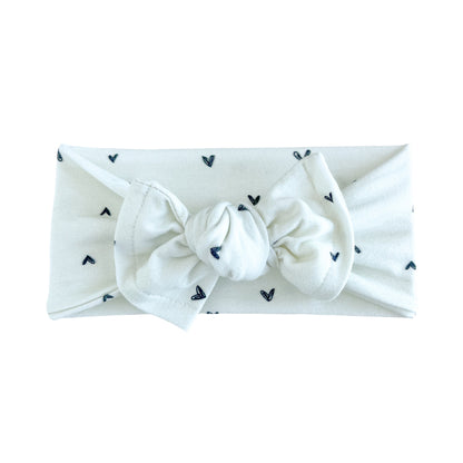 Top knot baby headband in off white with dainty black heart print, crafted from cotton jersey knit fabric, displayed on a white background