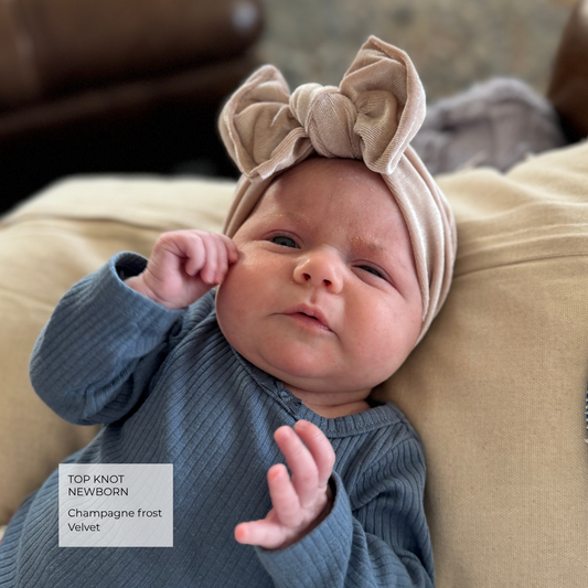 A newborn baby wearing a champagne beige top knot headband with a large bow and a ribbed blue-gray long-sleeve bodysuit, lying on a beige cushion. A text overlay in the bottom left reads 'TOP KNOT NEWBORN Champagne frost Velvet'.
