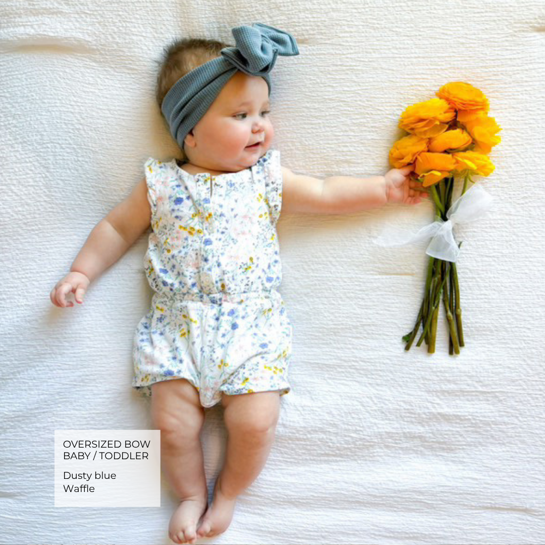 A baby lying on a white textured blanket, wearing a dusty blue oversized bow headband and a white sleeveless romper with a blue and yellow floral print. The baby is holding a bouquet of orange flowers.
