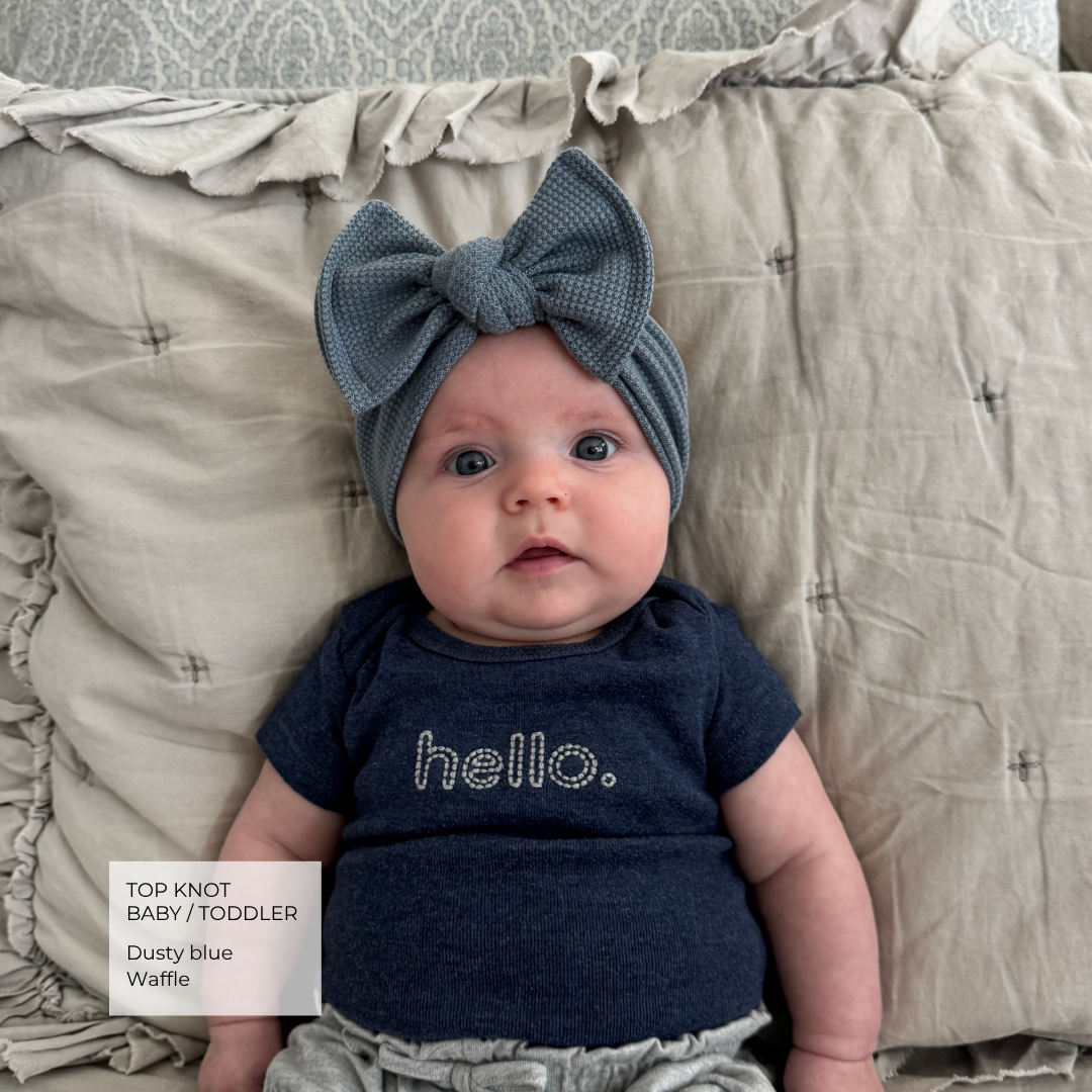 A baby lying on a beige quilted blanket, wearing a dusty blue textured top and a matching dusty blue top knot headband with a large bow. The shirt has the word 'hello.