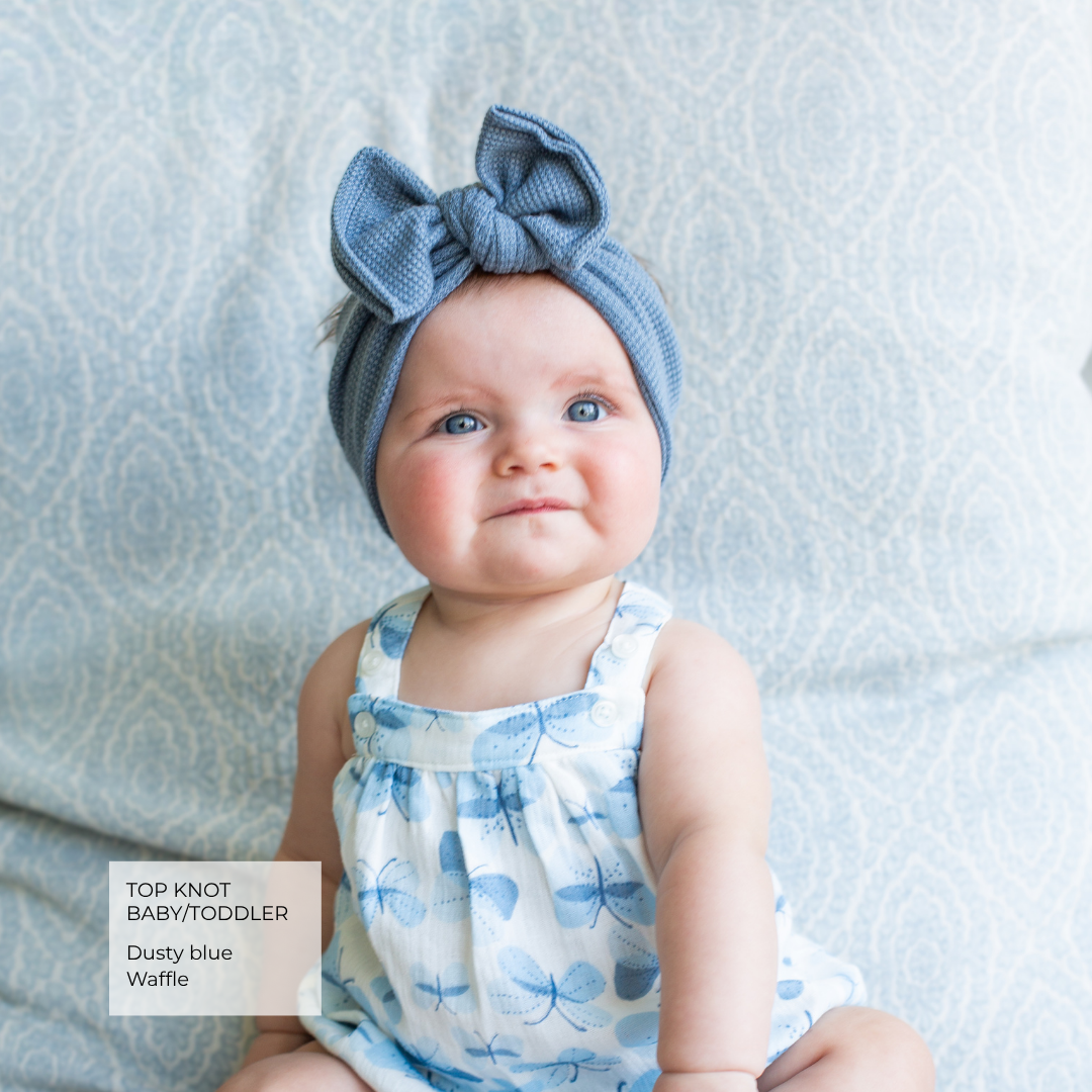 A baby wearing a dusty blue knotted top-knot baby/toddler headband and a matching blue-and-white butterfly print romper, sitting against a light blue patterned fabric background. A text box in the lower left reads 'TOP KNOT BABY/TODDLER Dusty blue Waffle'.